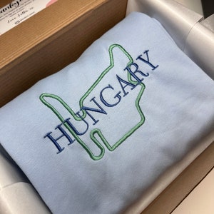 Puede incluir: Una sudadera de color azul claro con un dise&ntilde;o bordado en verde y azul de una mano haciendo el signo 'Te amo'. La palabra 'HUNGR&Iacute;A' est&aacute; bordada en azul sobre la mano.