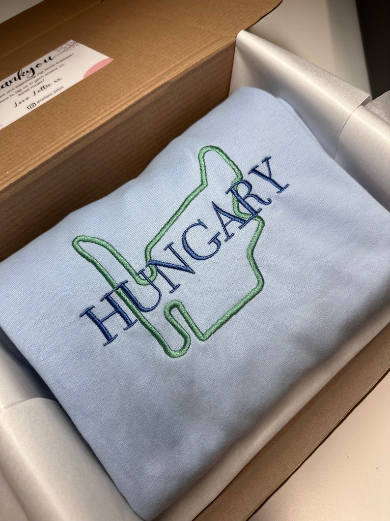 Puede incluir: Una sudadera de color azul claro con un dise&ntilde;o bordado en verde y azul de una mano haciendo el signo 'Te amo'. La palabra 'HUNGR&Iacute;A' est&aacute; bordada en azul sobre la mano.