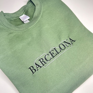 Puede incluir: Una sudadera verde con texto bordado en negro que dice "BARCELONA" en una fuente estilizada.