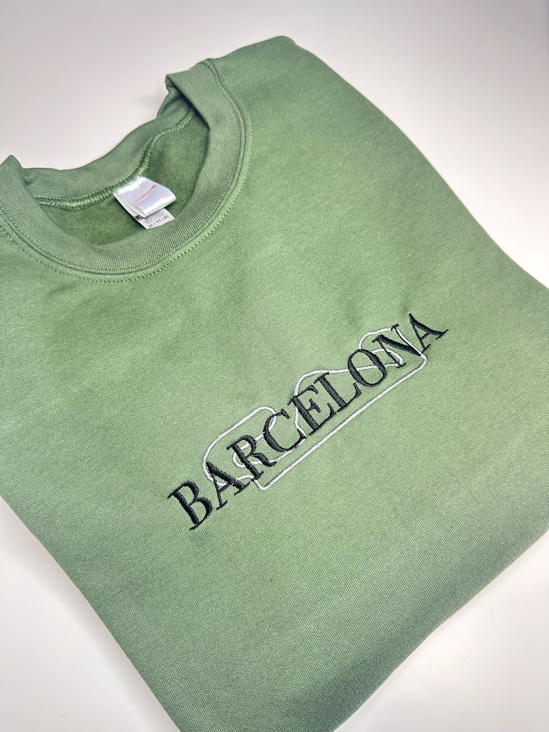 Puede incluir: Una sudadera verde con texto bordado en negro que dice "BARCELONA" en una fuente estilizada.