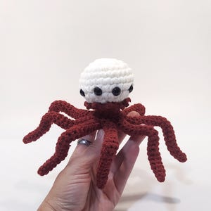 Op de afbeelding: Een handgemaakt gehaakt octopus speelgoed. De octopus heeft een witte kop met zwarte ogen en een bordeauxrode lichaam en tentakels. De tentakels zijn lang en gebogen. Het speelgoed is gemaakt van zacht garen.