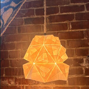 Puede incluir: Una lámpara colgante de papel geométrico, iluminada desde dentro, cuelga de una cuerda natural contra una pared de ladrillo. La luz tiene un brillo cálido y acogedor, y el papel está plegado en una forma compleja y multifacética.