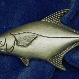 May include: A silver-colored pewter fish figurine with detailed scales and fins. The fish is lying on a blue velvet surface.
