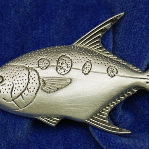 May include: A silver-colored pewter fish pin with a detailed design. The fish has a rounded body with a pointed head and a long, thin tail. The fish is covered in small dots and has several larger circles on its side.