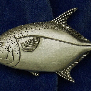 May include: A silver-colored pewter fish figurine with detailed scales and fins. The fish has a rounded body and a long, thin tail.