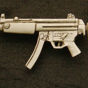 May include: A silver-colored miniature submachine gun, featuring detailed design elements such as a magazine, trigger, and sight. The small firearm is set against a dark background, highlighting its metallic finish and intricate construction.