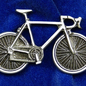 May include: A silver-colored bicycle pin with detailed spokes, chain, and handlebars. The bike is set against a deep blue background, highlighting the intricate design of the road bike. This pin is a great accessory for cyclists.