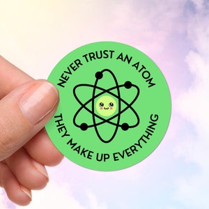 May include: A round, light green sticker with the text "NEVER TRUST AN ATOM THEY MAKE UP EVERYTHING" in black. The sticker features a cartoon atom with a smiling face. The sticker is held by a hand against a cloudy sky background.