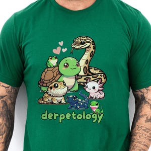 A man wearing a green t-shirt that features a colorful and adorable cartoon illustration of various reptiles and amphibians, including a snake, turtle, frog, axolotl, gecko, and salamander. Above the group are pink hearts, and below them is the playful word "derpetology," combining "derp" and "herpetology." The person has tattooed arms, is wearing light denim jeans, and is standing against a white wall with a green plant and a denim jacket in the background.