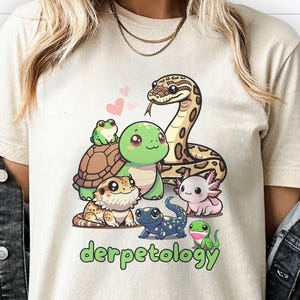K&ouml;nnte beinhalten: Ein cremefarbenes T-Shirt mit einer grafischen Illustration verschiedener Cartoon-Reptilien und Amphibien, darunter eine Schlange, eine Schildkr&ouml;te, ein Frosch, ein Axolotl und eine Eidechse, mit dem gr&uuml;nen Text "derpetology" darunter.