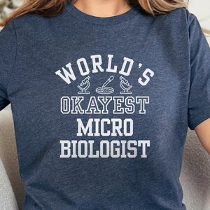May include: A heather navy blue short-sleeve t-shirt featuring white screen-printed text that reads "WORLD'S OKAYEST MICRO BIOLOGIST" with small graphic icons of two microscopes and a petri dish positioned above the lettering.
