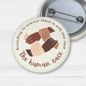 May include: Two round badges with a pin back. One badge displays a graphic of hands in various skin tones clasped together, with the text "According to science there is only one race, the human race."