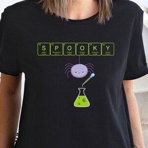 A black t-shirt displaying a science-themed Halloween design. The word "SPOOKY" is spelled using periodic table elements: Sulfur (S), Phosphorus (P), Oxygen (O), another Oxygen (O), Potassium (K), and Yttrium (Y), each with atomic data. Below the text, a smiling purple spider holds a pipette above a green bubbling beaker. The shirt is modeled indoors with Halloween decorations like striped books and black bat wall decals.