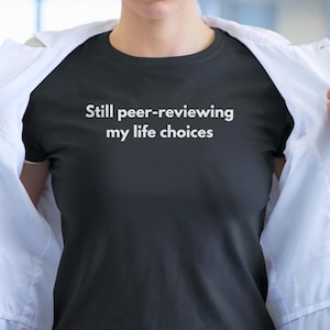 Può includere: Una maglietta nera con la scritta bianca "Still peer-reviewing my life choices". La maglietta viene mostrata da una persona che indossa un camice bianco.