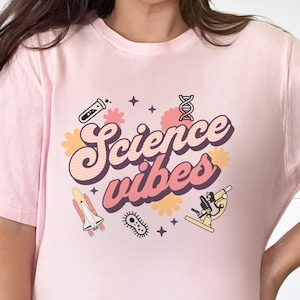 May include: Pale pink t-shirt with the text "Science Vibes" in a retro style. The design includes science-related illustrations such as a test tube, DNA helix, microscope, and a rocket. The graphics are in pink, yellow, and purple.