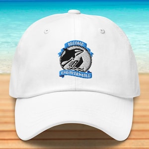 May include: White baseball cap with a black and blue embroidered design. The design features a whale, waves, and the words "BECOME UNGOVERNABLE" on a blue banner. The cap is on a wooden surface with a beach and ocean background.