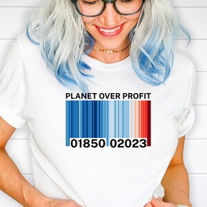 Może przedstawiać: Biała koszulka z napisem "PLANET OVER PROFIT" nad wzorem kodu kreskowego. Kod kreskowy przechodzi od niebieskiego do czerwonego, z numerami "01850 02023" poniżej. Codzienna odzież.