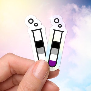 Puede incluir: Dos pegatinas con forma de tubos de ensayo, cada una con una combinación de colores diferente. Una presenta negro, gris y blanco, y la otra negro, gris, blanco y morado. Las pegatinas tienen un acabado holográfico y se sostienen en una mano.