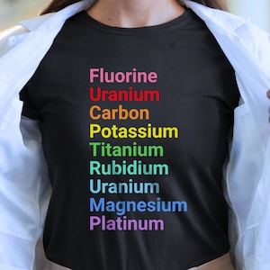 May include: Black t-shirt with a colorful periodic table of elements design. The elements listed are Fluorine, Uranium, Carbon, Potassium, Titanium, Rubidium, Uranium, Magnesium, and Platinum. The text is in a rainbow of colors.
