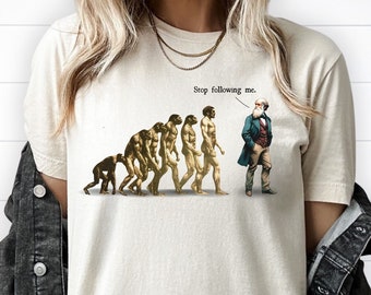 May include: A beige t-shirt featuring a graphic of the evolution of humans, culminating in Charles Darwin. The text "Stop following me." is positioned above Darwin's head.