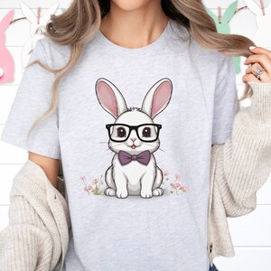 May include: Light gray t-shirt featuring a cartoon bunny wearing glasses and a bow tie. The bunny is white with pink ears and is sitting in front of small pink flowers. Decorative bunny cutouts hang in the background.