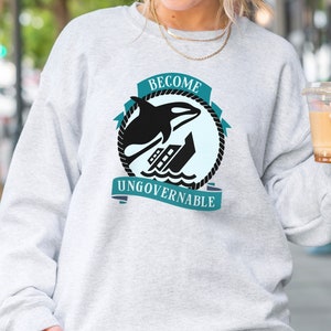 May include: A grey sweatshirt with a graphic of a killer whale jumping over a ship. The graphic is surrounded by a blue rope and the text "Become Ungovernable".