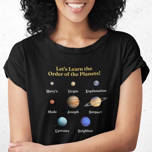 May include: A black t-shirt with a yellow graphic that says "Let's Learn the Order of the Planets!" and features a mnemonic device for remembering the order of the planets in our solar system. The planets are represented by images and each image has a word underneath it. The words are: "Mary's", "Virgin", "Explanation", "Made", "Joseph", "Suspect", "Upstairs", "Neighbor".