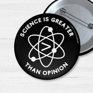 May include: A round black pin-back button featuring a white atom graphic at the centre with a greater-than symbol inside. The text "SCIENCE IS GREATER THAN OPINION" is printed in white capital letters in a circular arc around the atom graphic.