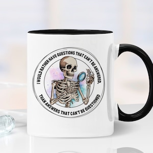 May include: White coffee mug with a black handle and black rim. The mug features a black and white illustration of a skeleton holding a magnifying glass. The text on the mug reads "I would rather have questions that can't be answered, than answers that can't be questioned."