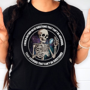 May include: Black t-shirt with a white circle graphic featuring a skeleton holding a magnifying glass in front of a galaxy background. The text around the graphic reads "I would rather have questions that can't be answered, than answers that can't be questioned."