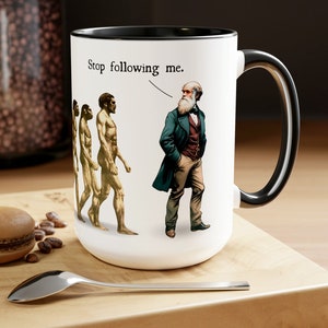 May include: White ceramic mug with a black handle and rim. The mug features a humorous illustration of the theory of evolution with a caption that reads "Stop following me."
