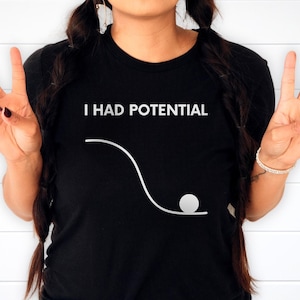 May include: Black t-shirt with the white text "I HAD POTENTIAL" above a white graphic of a ball rolling down a curved path. The person is making a peace sign with their fingers.