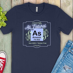 May include: A navy blue t-shirt with a white graphic of a periodic table element. The element is arsenic, with the symbol "As", atomic number 33, and atomic weight 74.922. The graphic is surrounded by a decorative border and the text "100% All Natural, Non-GMO, Gluten Free".
