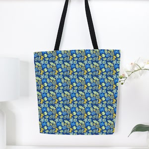 May include: A blue tote bag featuring a repeating pattern of abstract shapes in blue and yellow. The bag has black handles and is suspended against a white wall. A reusable shopping bag.