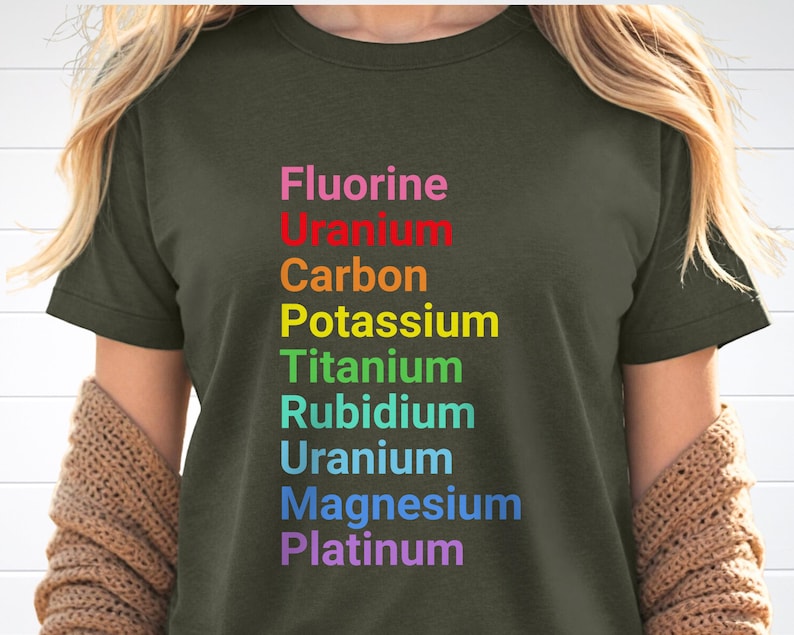 May include: An olive green t-shirt featuring a vertical list of chemical elements printed in a rainbow color gradient. The text reads: Fluorine, Uranium, Carbon, Potassium, Titanium, Rubidium, Uranium, Magnesium, and Platinum in stacked rows.