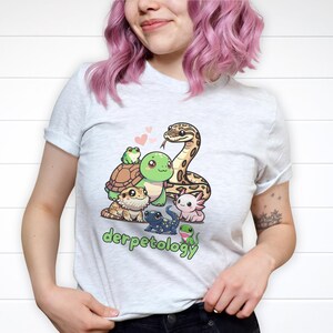 K&ouml;nnte beinhalten: Ein hellgraues T-Shirt mit einer bunten Grafik verschiedener Cartoon-Reptilien und Amphibien, darunter eine Schlange, eine Schildkr&ouml;te, ein Frosch und ein Axolotl, mit dem Schriftzug "derpetology" in gr&uuml;nen Buchstaben unter der Illustration.