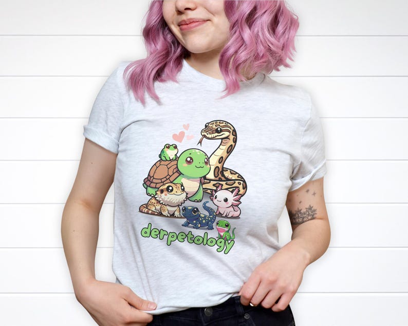 K&ouml;nnte beinhalten: Ein hellgraues T-Shirt mit einer bunten Grafik verschiedener Cartoon-Reptilien und Amphibien, darunter eine Schlange, eine Schildkr&ouml;te, ein Frosch und ein Axolotl, mit dem Schriftzug "derpetology" in gr&uuml;nen Buchstaben unter der Illustration.