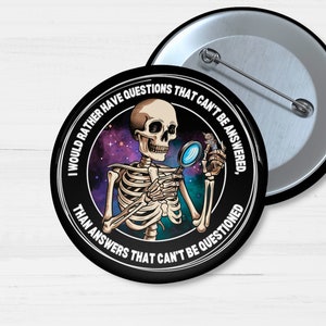 May include: A black and white button with a skeleton holding a magnifying glass. The text on the button reads "I would rather have questions that can't be answered, than answers that can't be questioned."