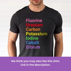 May include: A charcoal gray short-sleeve t-shirt featuring a vertical list of chemical elements printed in rainbow colors: Fluorine in pink, Uranium in red, Carbon in orange, Potassium in yellow, Iodine in green, Cobalt in blue, and Erbium in purple.