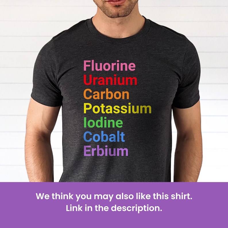May include: A charcoal gray short-sleeve t-shirt featuring a vertical list of chemical elements printed in rainbow colors: Fluorine in pink, Uranium in red, Carbon in orange, Potassium in yellow, Iodine in green, Cobalt in blue, and Erbium in purple.