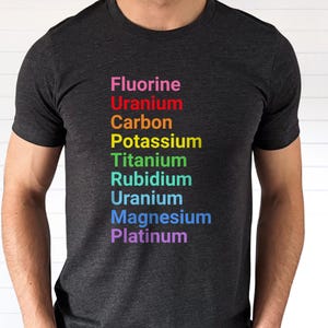 May include: A dark charcoal gray short-sleeve t-shirt featuring a vertical list of chemical elements printed in rainbow colors. The text reads Fluorine, Uranium, Carbon, Potassium, Titanium, Rubidium, Uranium, Magnesium, and Platinum in sequence.