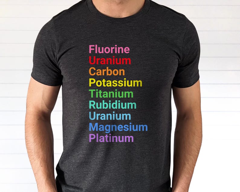 May include: A dark charcoal gray short-sleeve t-shirt featuring a vertical list of chemical elements printed in rainbow colors. The text reads Fluorine, Uranium, Carbon, Potassium, Titanium, Rubidium, Uranium, Magnesium, and Platinum in sequence.