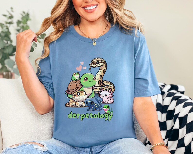 K&ouml;nnte beinhalten: Ein blaues Kurzarm-T-Shirt mit einem Grafikdruck verschiedener Reptilien und Amphibien, darunter eine Schlange, eine Schildkr&ouml;te, ein Frosch, ein Axolotl und eine Eidechse, mit dem gr&uuml;nen Text "derpetology" und kleinen rosa Herzen.