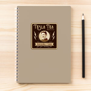 May include: A spiral-bound notebook with a tan cover featuring a Tesla Tea label. The label has a vintage design with an image of Nikola Tesla, lightning bolts, and the text "Shockingly Good". A black pencil rests beside the notebook.