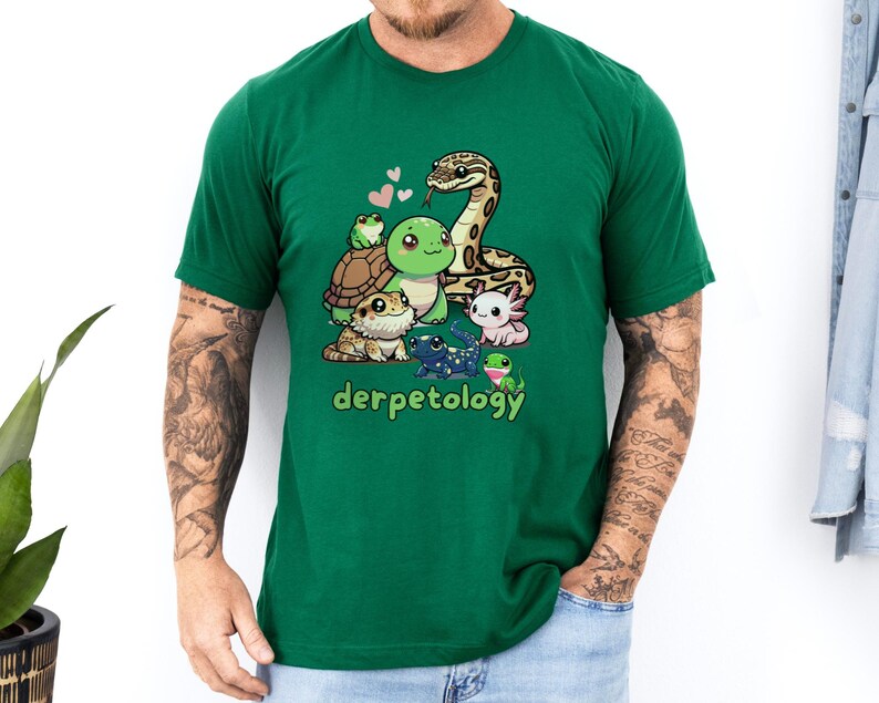 A man wearing a green t-shirt that features a colorful and adorable cartoon illustration of various reptiles and amphibians, including a snake, turtle, frog, axolotl, gecko, and salamander. Above the group are pink hearts, and below them is the playful word "derpetology," combining "derp" and "herpetology." The person has tattooed arms, is wearing light denim jeans, and is standing against a white wall with a green plant and a denim jacket in the background.