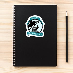 May include: A black notebook with a spiral binding and a sticker on the front. The sticker is teal blue and white and features a black orca jumping over a ship. The sticker says "Become Ungovernable".