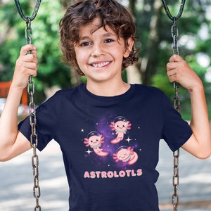 K&ouml;nnte beinhalten: Ein marineblaues T-Shirt mit einem Weltraum-Design, das rosa Axolotl zeigt, die Astronautenhelme tragen. Der Text "ASTROLOTLS" ist in rosa Buchstaben auf dem Shirt gedruckt.
