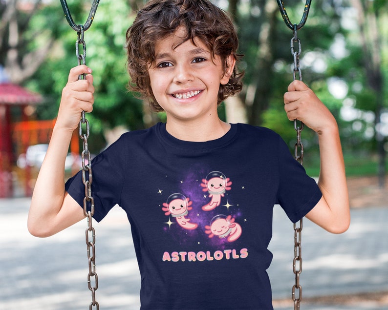 K&ouml;nnte beinhalten: Ein marineblaues T-Shirt mit einem Weltraum-Design, das rosa Axolotl zeigt, die Astronautenhelme tragen. Der Text "ASTROLOTLS" ist in rosa Buchstaben auf dem Shirt gedruckt.