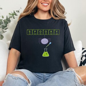 May include: Black t-shirt with the word "SPOOKY" spelled out using elements from the periodic table. A purple spider, a beaker, and a pipette are featured in the design. The shirt is a Halloween-themed graphic tee.