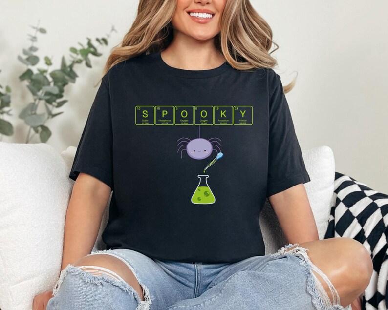 May include: Black t-shirt with the word "SPOOKY" spelled out using elements from the periodic table. A purple spider, a beaker, and a pipette are featured in the design. The shirt is a Halloween-themed graphic tee.